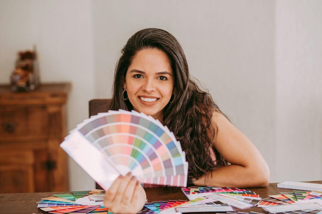 A woman with long hair is sitting at a table holding color swatches, smiling warmly.